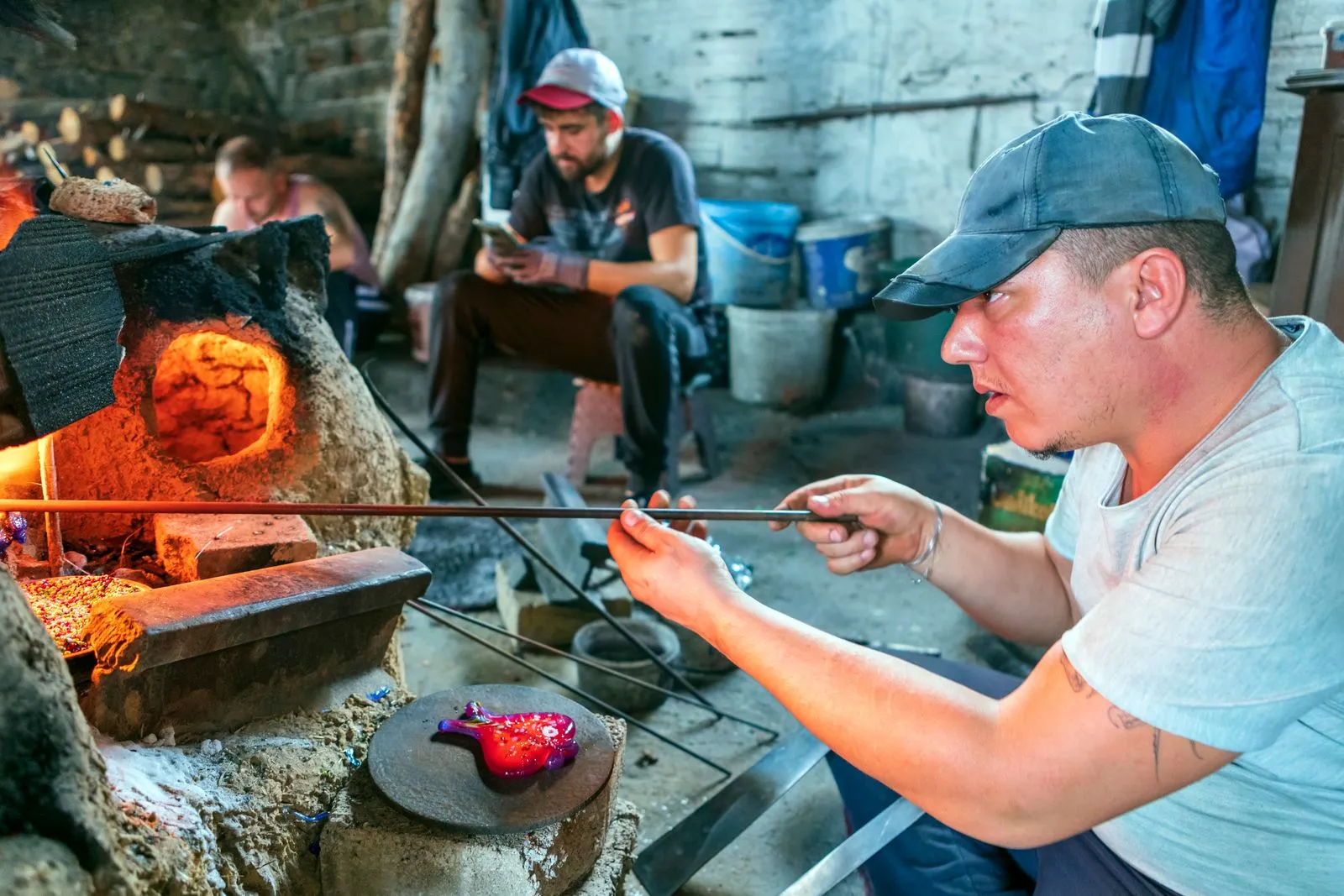 A glassblower works molten glass on a rod beside a glowing furnace in a workshop.