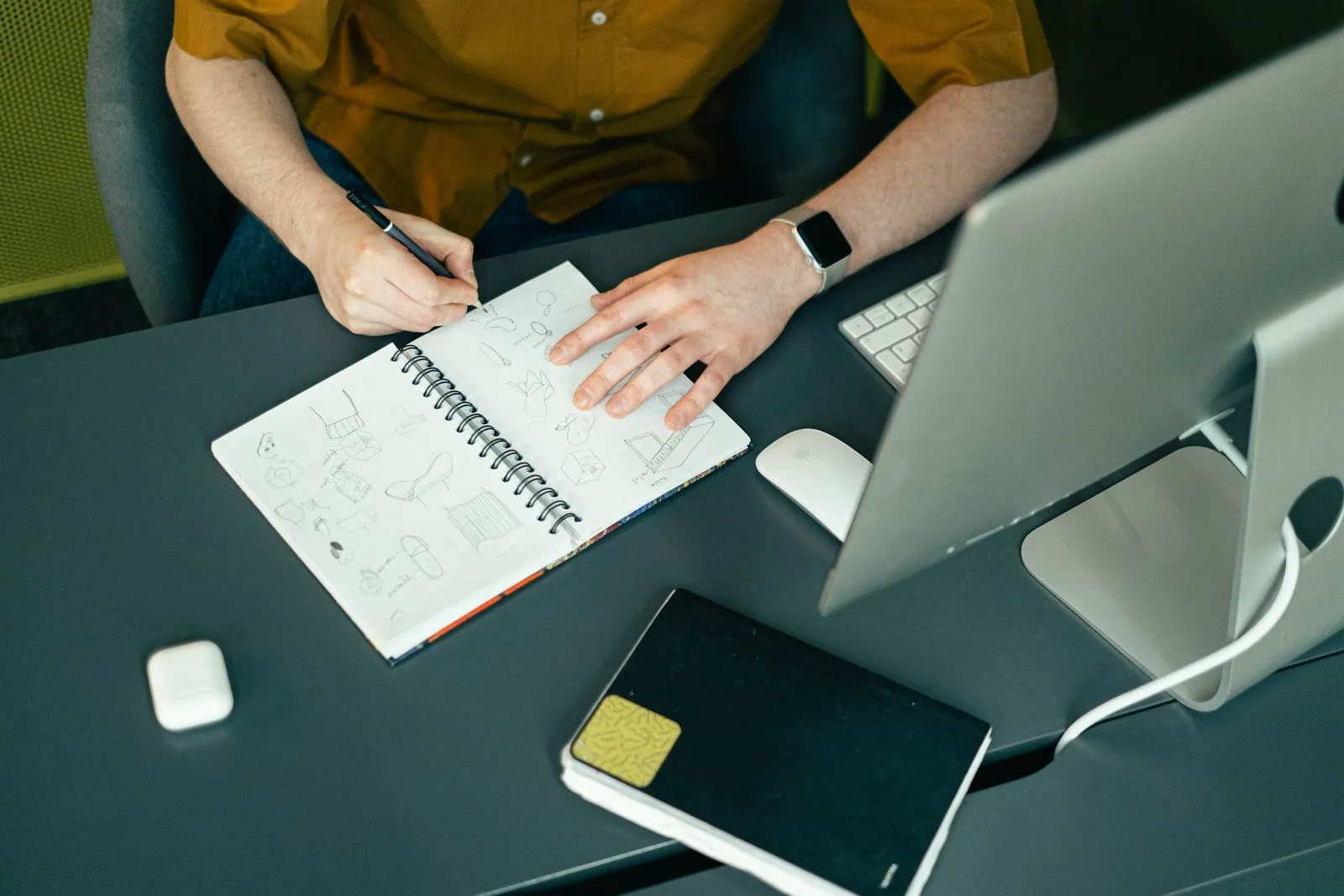 Close-up of a hand sketching object designs in a spiral notebook next to a desktop computer.