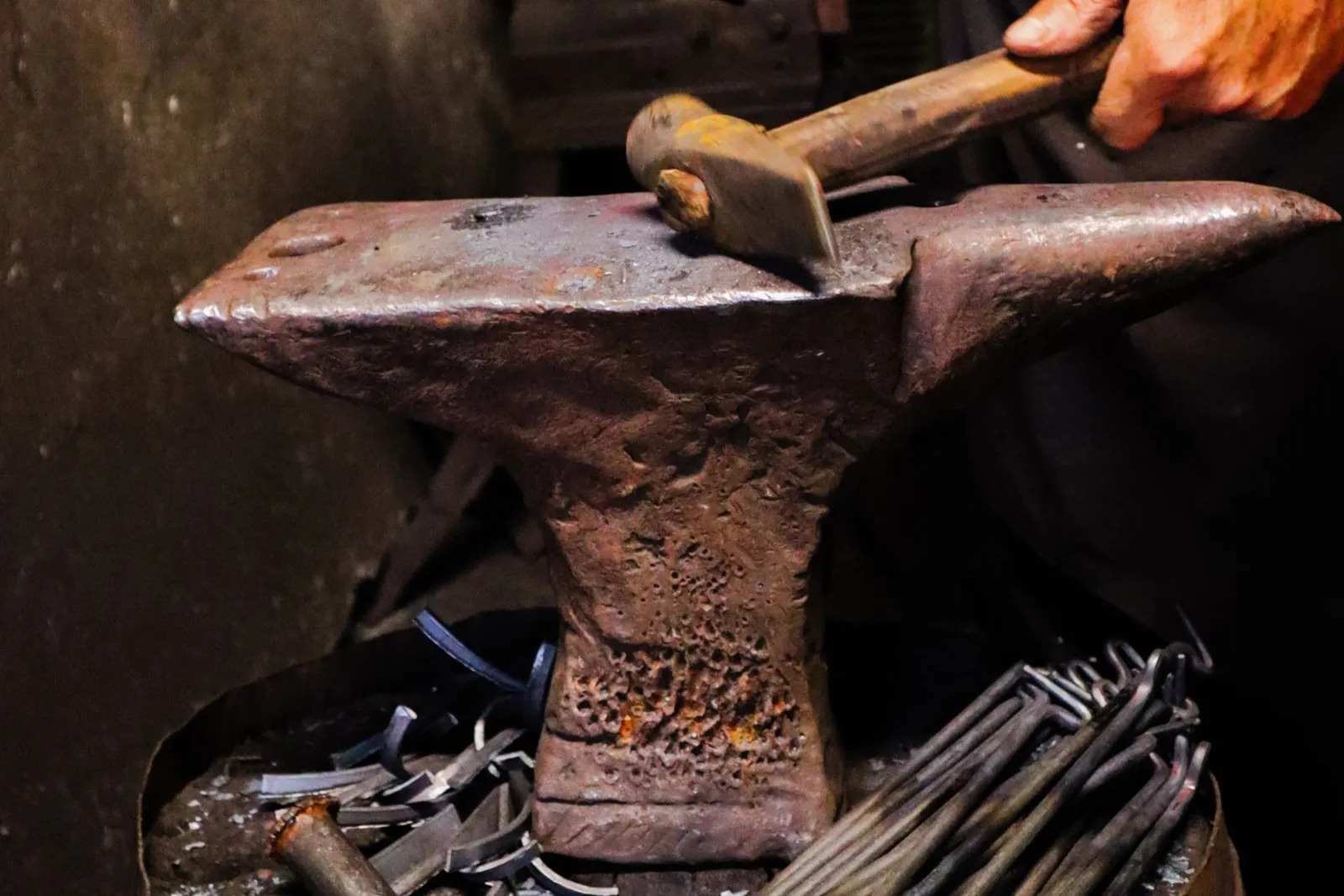 A blacksmith's hammer strikes glowing iron on an anvil, with tongs and forged pieces nearby.