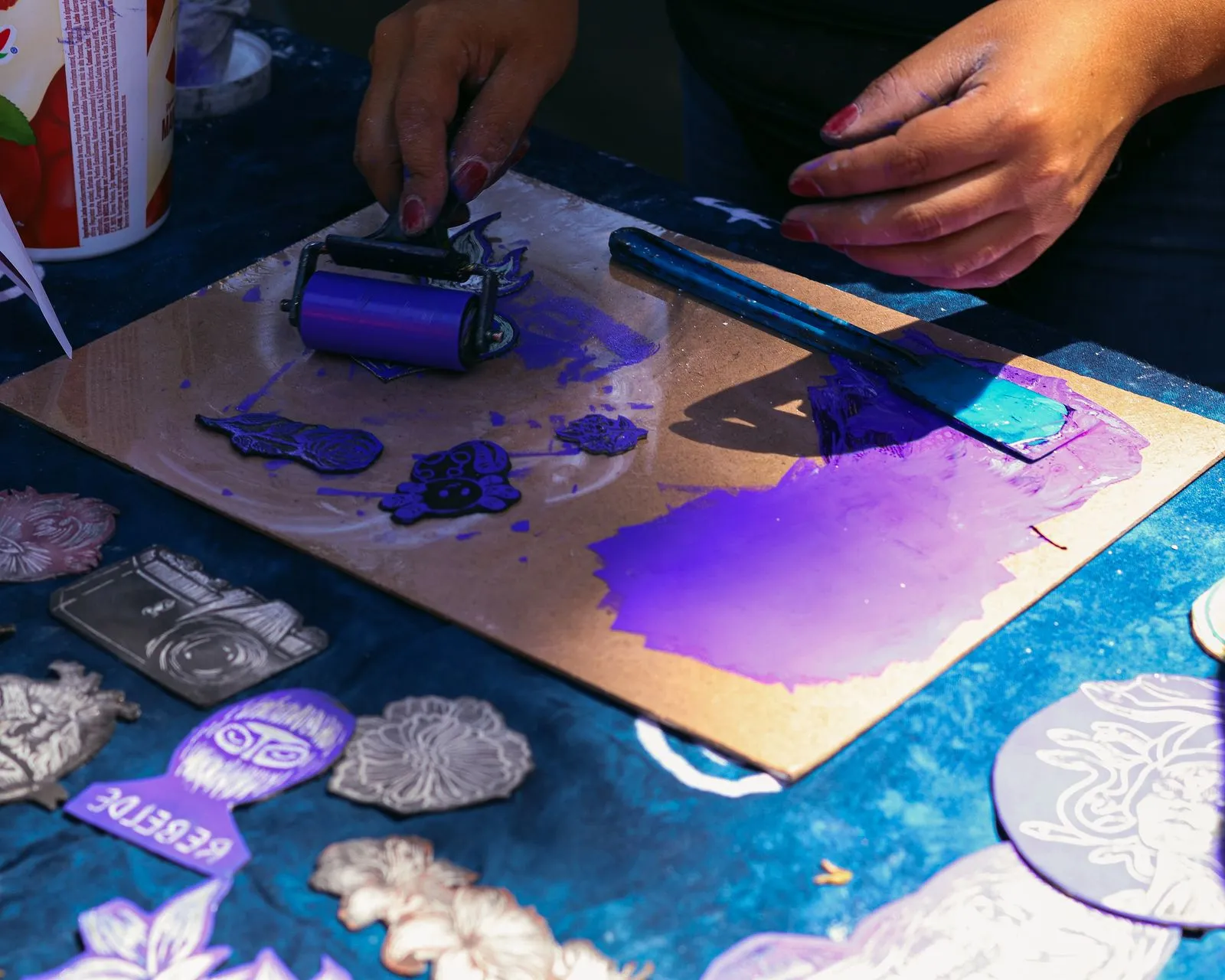 Hands roll purple ink onto a carved linocut printing block surrounded by finished relief prints.