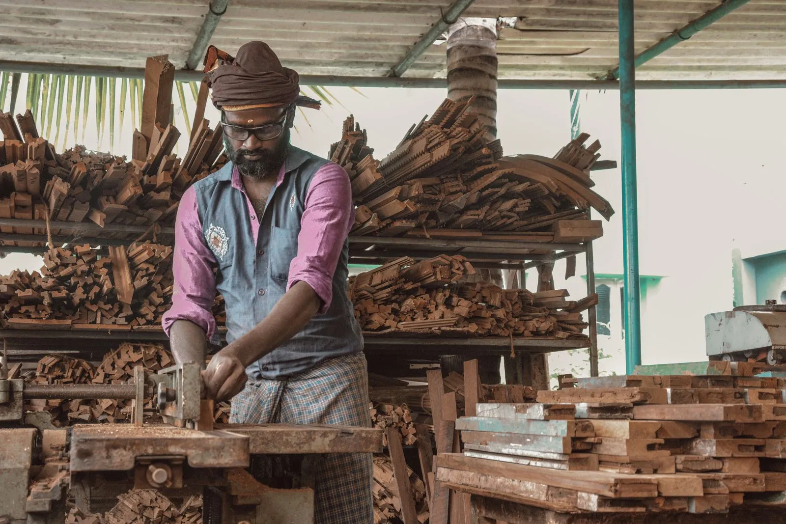A woodworker sands a piece of furniture by hand in a workshop surrounded by stacked lumber.