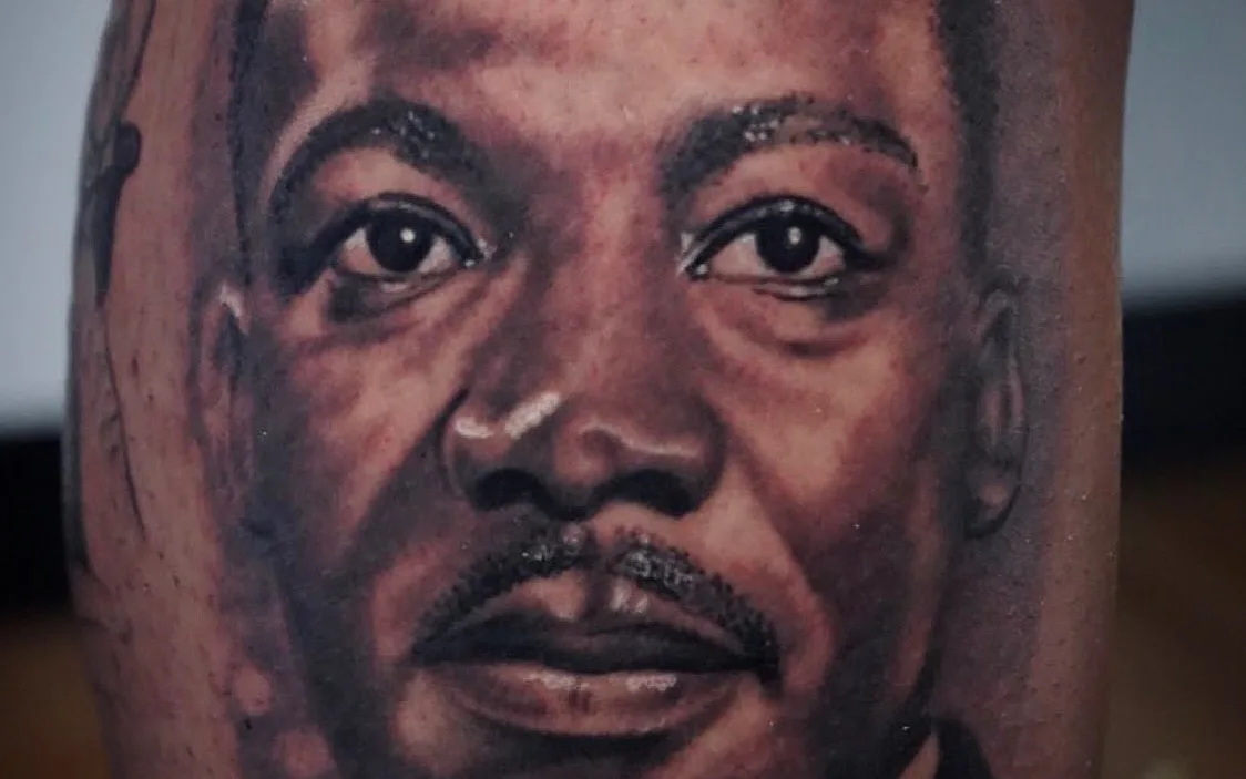 A black-and-grey realism portrait tattoo of Dr. Martin Luther King Jr. on a client's forearm, cropped to the tattoo only.